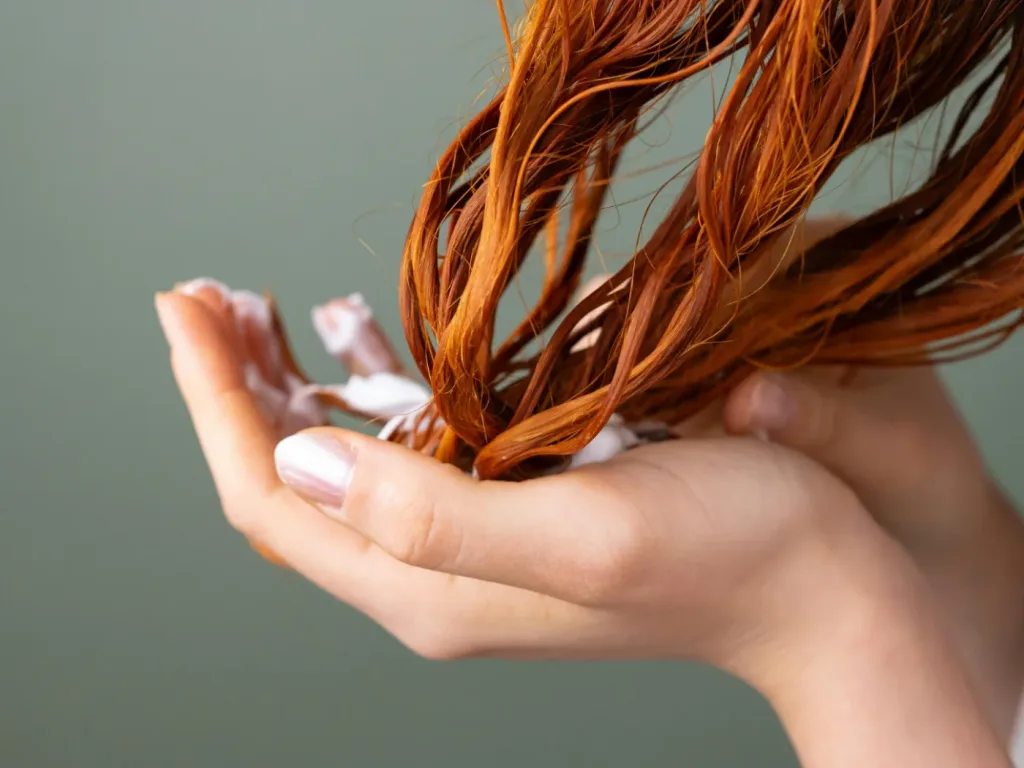 Mano sosteniendo un mechón de cabello pelirrojo húmedo sobre un fondo verde apagado.