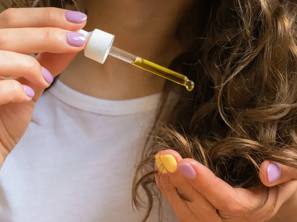 Manos aplicando un producto con cuentagotas sobre cabello rizado castaño claro.